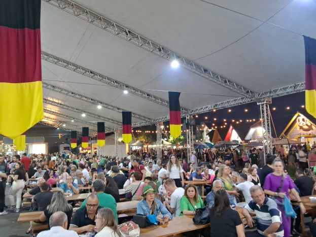 Große Gruppe von Menschen an langen Tischen unter einem Zelt auf dem Oktoberfest in München, Deutschland, mit Taschen und Gegenständen auf den Tischen und festlicher Décoration im Hintergrund.