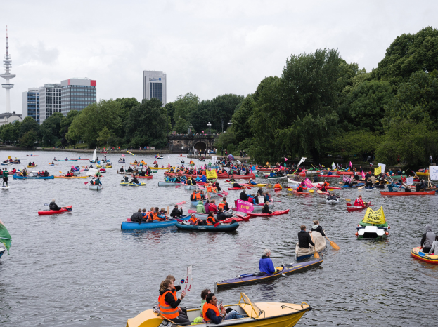 Eine große Gruppe von Menschen in Kanus auf einem Gewässer, einige halten Paddel und Fahnen, mit Bäumen, Gebäuden und einem Turm im Hintergrund.