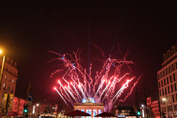 Eine belebte Stadtstraße an einem Silvesterabend in Berlin, voller Menschen, Fahrzeuge und Gebäude, beleuchtet von Straßenlaternen, Gebäudelichtern und Feuerwerk am Himmel.