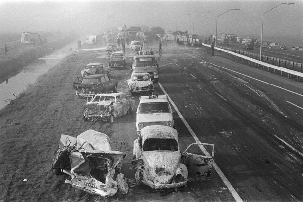 Ein Schwarz-Weiß-Foto von mehreren verunglützten Autos am Straßenrand bei Nacht, mit Laternen, nahestehenden Menschen, Bäumen und einem Gewässer im Hintergrund.