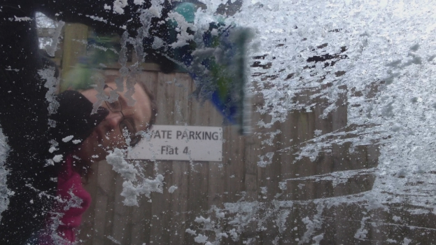 Ein mit Schnee gefülltes Glas ist im Vordergrund zu sehen, im Hintergrund eine Person und eine hölzerne Wand, auf der ein Schild mit Text angebracht ist.