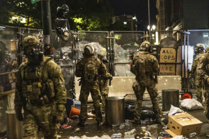 Eine Gruppe von Polizisten in Uniform und Helmen steht nachts vor einem Zaun, hält Waffen in den Händen und hat Müll und Trümmer auf dem Boden verstreut, sowie Gebäude, Bäume und ein Schild im Hintergrund.