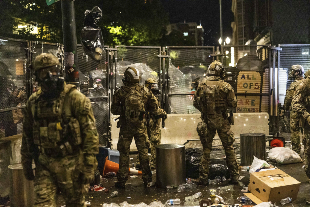Eine Gruppe von Polizisten in Uniform und Helmen steht nachts vor einem Zaun, hält Waffen in den Händen und hat Müll und Trümmer auf dem Boden verstreut, sowie Gebäude, Bäume und ein Schild im Hintergrund.