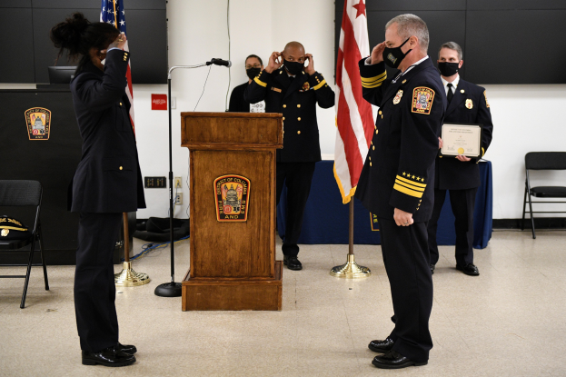 Eine Gruppe uniformierter Personen in Masken steht vor einem Podium mit Mikrofon, umgeben von Fahnen, Stühlen und Tischen, mit einer Person, die ein Zertifikat hält, während einer Vereidungszeremonie für einen neuen Feuerchef.