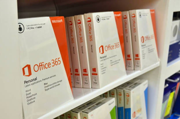Ein weißes Regal mit ordentlich angeordneten Microsoft Office 365-Schachteln, auf denen das Microsoft-Logo deutlich zu sehen ist, vor einem leicht unscharfen Hintergrund.