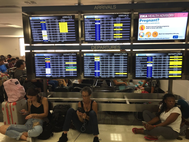 Gruppe von Menschen, die auf dem Boden eines Flughafens sitzen, einige halten Handys, mit verstreuten Taschen und Bildschirmen, die Zika-Virus-Informationen anzeigen.