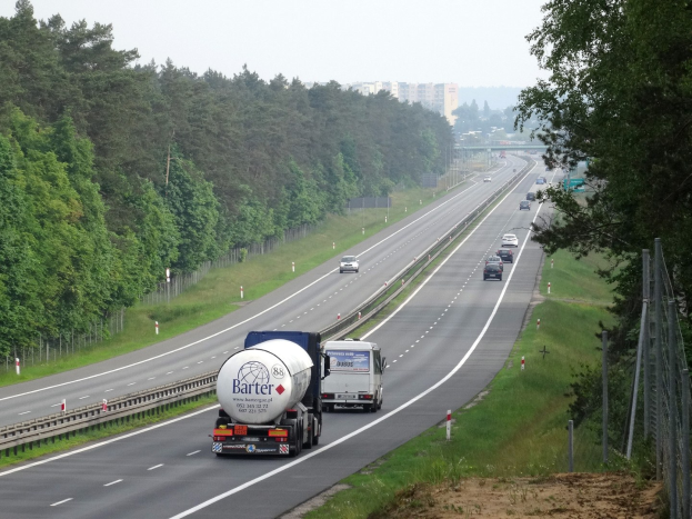 Ein großer Lkw fährt auf einer von beiden Seiten von Bäumen gesäumten Autobahn, mit einem Zaun und Gras auf der rechten Seite und Gebäuden in der Ferne unter einem klaren blauen Himmel.