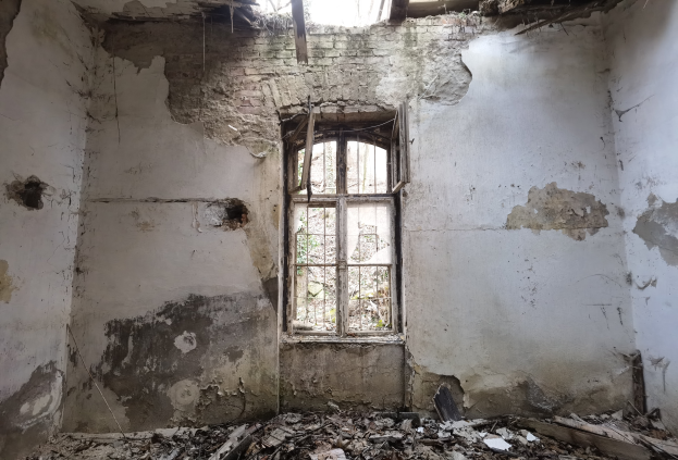 Ein alter Raum mit abblätternden Wänden, ein mittiges Fenster und ein Haufen Schrott auf dem Boden, der auf Verlassheit hindeutet.