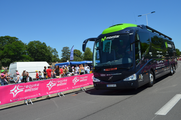 Ein schwarzer und grüner Bus fährt auf einer Straße neben einer Menschenmenge, einige tragen Mützen, mit einem Banner auf der linken Seite und Bäumen unter einem klaren blauen Himmel im Hintergrund.