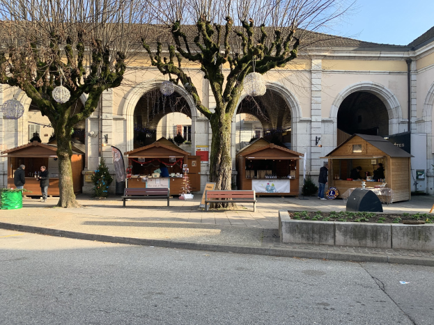 Ein belebter Weihnachtsmarkt mit Ständen und Menschen vor einem Gebäude mit Bögen und Säulen, umgeben von Bäumen und Bänken unter einem sichtbaren Himmel.
