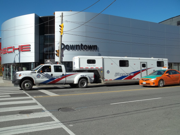 Ein Polizeilaster ist vor einem Gebäude mit Text darauf geparkt, mit einer Verkehrsampel mit einem Mast und Drähten im Vordergrund und Bäumen und einem klaren blauen Himmel im Hintergrund.