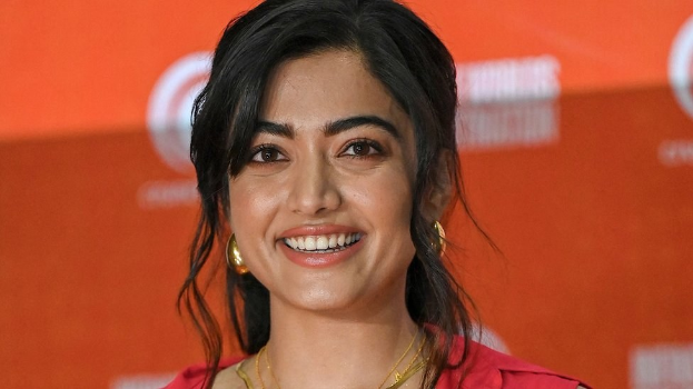 Eine Frau in einem roten Kleid mit einem eleganten Dutt und dezentem Make-up steht lächelnd da, trägt schwarze High Heels und hat ein hellgelbes Banner mit der Aufschrift "Sonam Kapoors beste Schönheits Looks vom Cannes Film Festival" im Hintergrund.