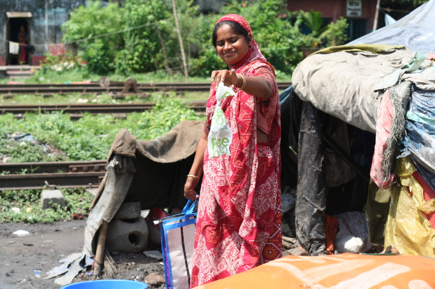 Eine Frau in einem roten Sari lächelt, hält eine weiße Decke in der Hand und steht neben einem kleinen blauen Zelt mit Eisenbahnschienen und Bäumen im Hintergrund.