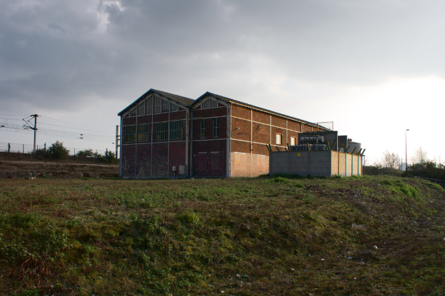 Eine Szene mit Gartenhütten, Transformatoren, Strommasten mit Kabeln, Zäunen, einer Straßenlaterne, Bäumen und einem bewölkten Himmel.