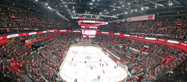 Eine große Indoor-Eishockeyarena mit Zuschauern auf den Rängen, heller Deckenbeleuchtung und einem zentralen Scoreboard-Bildschirm während eines Spiels.
