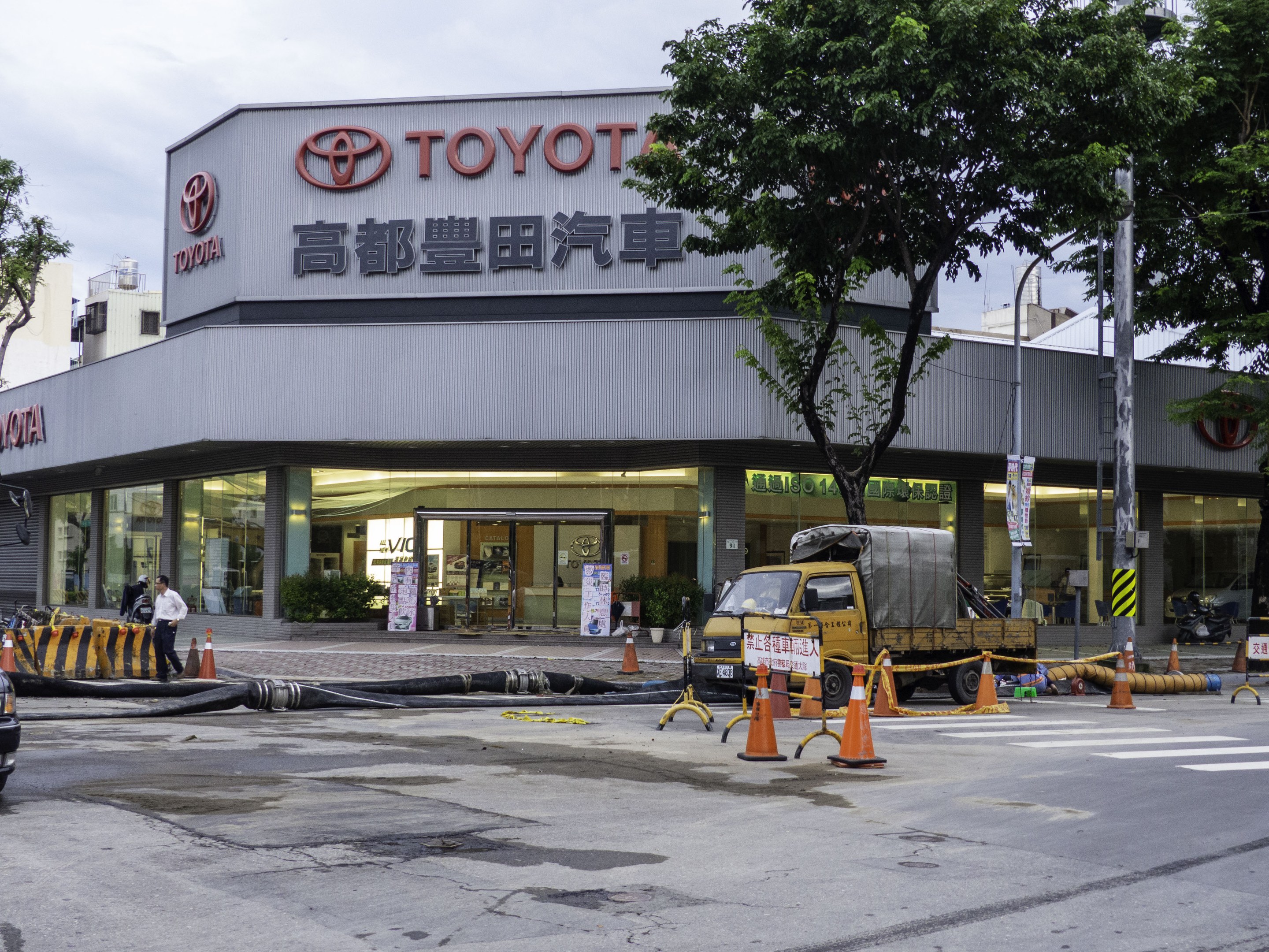 Ein Toyota-Autohaus in Tokio, Japan, mit Fahrzeugen, Verkehrskegeln, Rohren, Menschen, Bäumen, Polen, Brettern und Gebäuden im Vordergrund und einem bewölkten Himmel im Hintergrund.