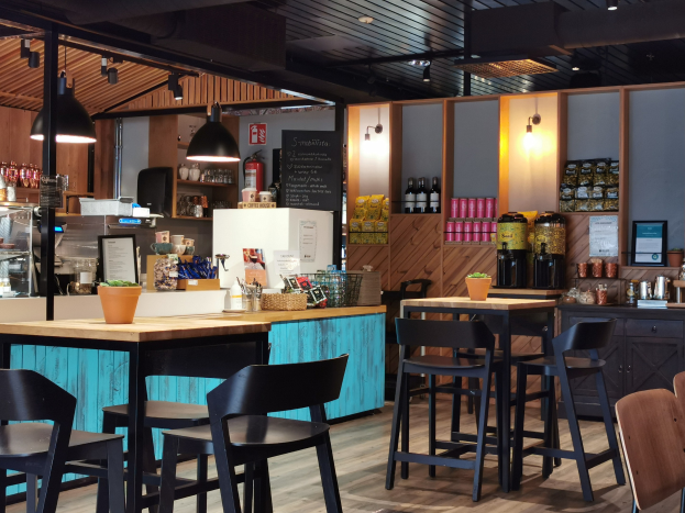 Innenraum eines Restaurants mit Holztischen, Stühlen, einer Topfpflanze auf einem Tisch, einer Schautafel, einem Kühlschrank, einem Feuerlöscher, Regalen mit Flaschen, Schränken mit Gegenständen, einem Foto auf der Wand, Deckenlampen und Lampen.