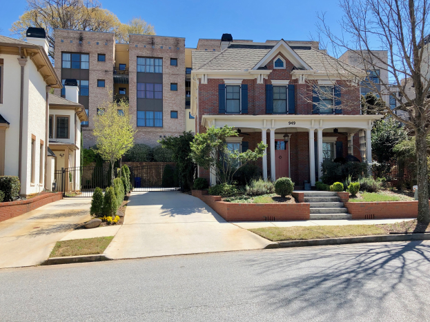 Eine Reihe von Stadth├Ąusern in einem Wohnviertel mit Einfahrten, umgeben von Pflanzen, Gras, B├Ąumen, einer Treppe, einem Metallzaun und einem Weg, unter einem bew├Âlktem Himmel.