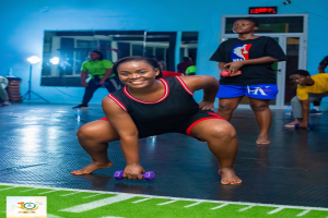 Eine Frau in einem Fitnessstudio führt eine Kniebeuge aus, während sie Hanteln hält, mit verschiedenen Fitnessgeräten und einem Bildschirm im Hintergrund, und Logos und Text am unteren Bildrand.