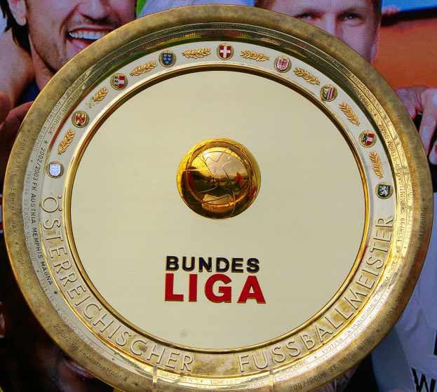 Ein Mann hält ein goldenes Schild mit der Aufschrift 'Bundesliga' hoch, im Hintergrund eine Fahne mit drei Personen.