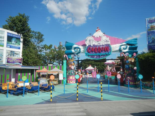 Karussell am Parkeingang mit Straße, eingezäuntem Bereich, Buden, Schildern, Bäumen und bewölktem Himmel im Hintergrund.