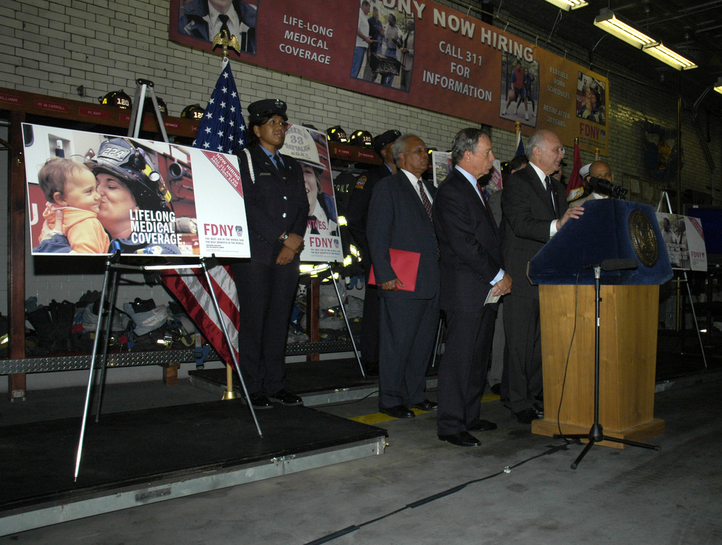 Eine Gruppe von Menschen steht in der Nähe eines Podiums mit Mikrofonen, Schildern auf Ständern, Fahnen und Schildern an der Wand, alles unter hellem Licht.