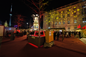 Ein lebendiger Weihnachtsmarkt in Berlin, Deutschland, mit Menschen um dekorierte Stände, Bäume, Gebäude, Laternenmasten und einen Turm unter einem dunklen Himmel.