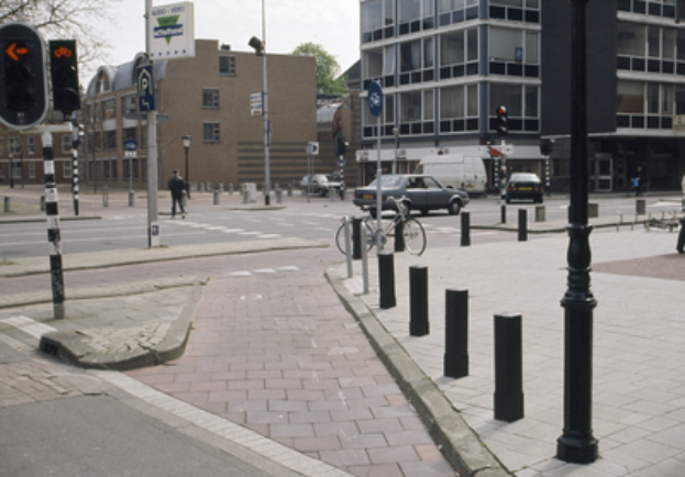 Eine Straßenszene in der Stadt mit einem geparkten Fahrrad, fahrenden Fahrzeugen, Fußgängern, Pfählen, Verkehrsampeln, Schildern, Bäumen, Gebäuden mit Fenstern und einem klaren blauen Himmel.