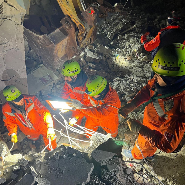 Eine Gruppe von Menschen in orangefarbener Uniform und grünen Helmen bei der Arbeit an Schutt mit einem Kran im Hintergrund bei einem Einsatz zur Bergung eines eingestürzten Gebäudes.