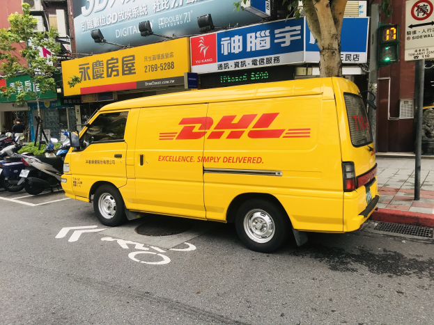 Ein gelber DHL-Lieferwagen parkt auf einer Straße neben Gebäuden, Bäumen, Schildern, Masten, Ampeln und einer Person auf dem Gehweg.