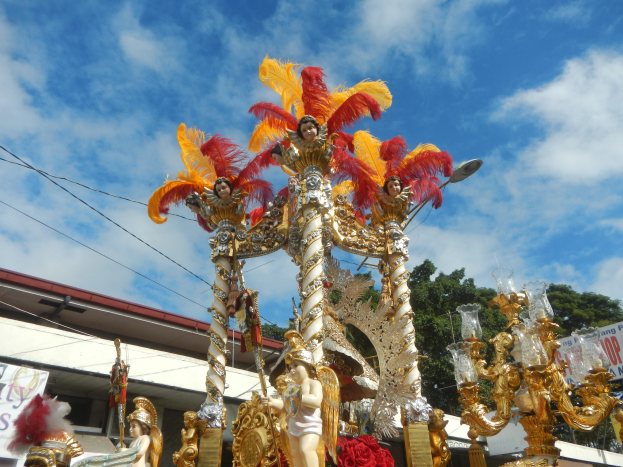 Ein Karnevalswagen mit goldenen und roten Federn, auf dem Menschen sitzen, wird in einem Umzug mit einem Gebäude, Bäumen, Drähten und einer Fahne in der Nähe sowie einem sichtbaren Himmel gezeigt.