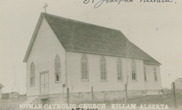 Ein Schwarz-Weiß-Foto von St. Joseph's Lutheran Church in Killam, Alberta, das ein Haus mit Dach, Fenstern, Türen und einem Kreuz auf dem Dach zeigt, umgeben von einem Zaun, Gras und anderen Häusern im Hintergrund, mit Text oben und unten im Bild.