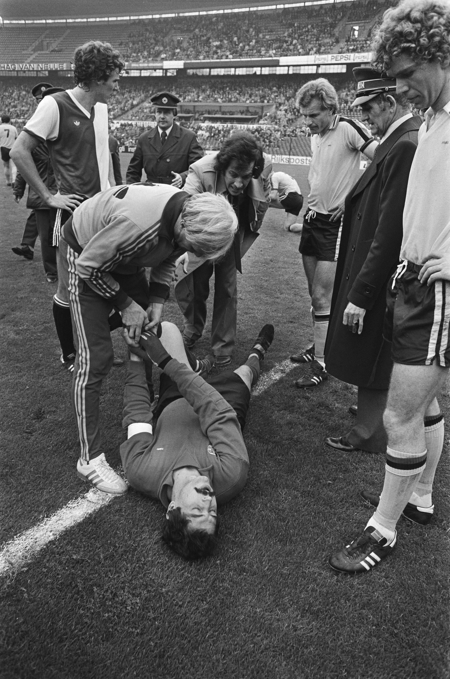 Schwarzes und weißes Bild eines Fußballspielers, der auf dem Boden liegt und von einer Gruppe Männer umgeben ist, mit einer Menschenmenge im Hintergrund eines Stadions.