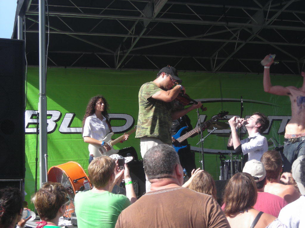 Eine Gruppe von Musikern auf der Bühne mit verschiedenen Instrumenten, während das Publikum davor steht und die Musik genießt, mit einem großen schwarzen Lautsprecher links und einem grünen Banner im Hintergrund.