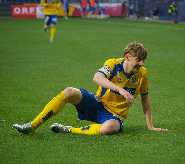 Ein Fußballspieler sitzt auf dem Boden und hat seinen Kopf in den Händen, umgeben von einer Gruppe von Menschen in Sportbekleidung, mit unscharfen Brettern, Geländern und Pfosten im Hintergrund.