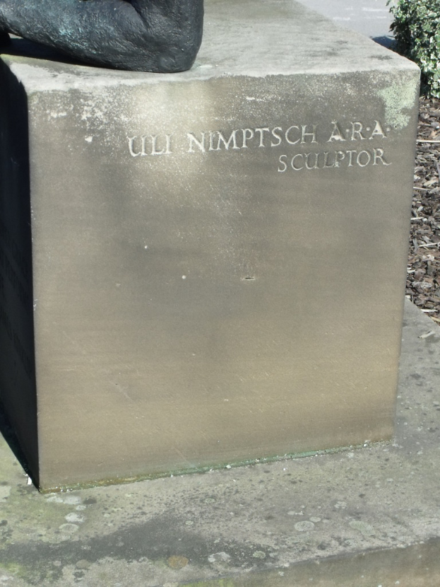 Ein Stein mit Text in Vordergrund, möglicherweise eine Statue oben, mit trockenen Zweigen, Erde und Pflanzen rechts und einer Straße im Hintergrund.