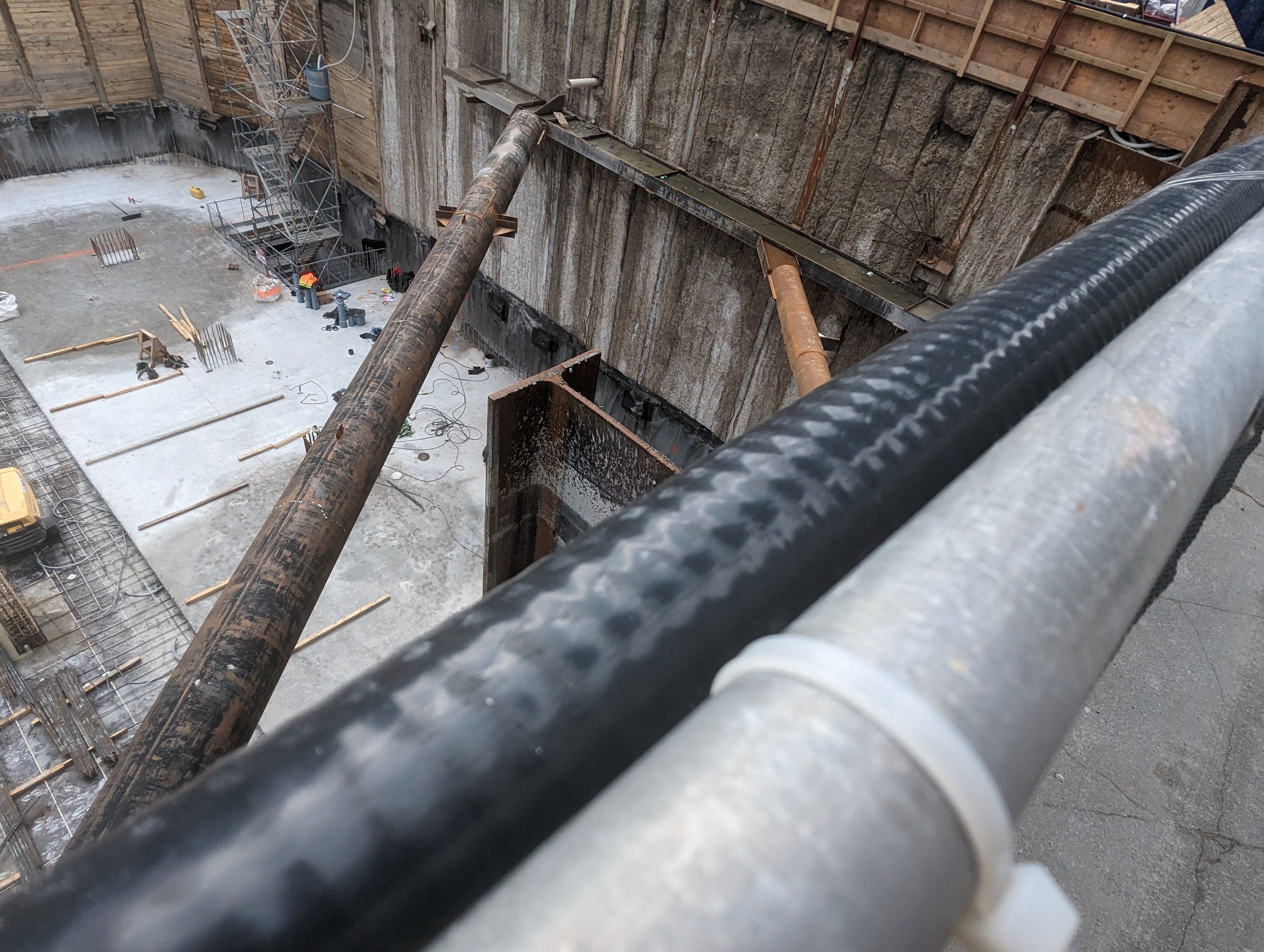 Großes Metallrohr auf einer Baustelle umgeben von Arbeitern, Holzwerkstoffen und Ausrüstung, mit einer Person in einem blauen Hemd rechts und Fahrzeugen und einem Gebäude im Hintergrund.