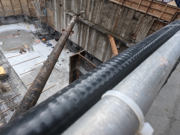 Großes Metallrohr auf einer Baustelle umgeben von Arbeitern, Holzwerkstoffen und Ausrüstung, mit einer Person in einem blauen Hemd rechts und Fahrzeugen und einem Gebäude im Hintergrund.