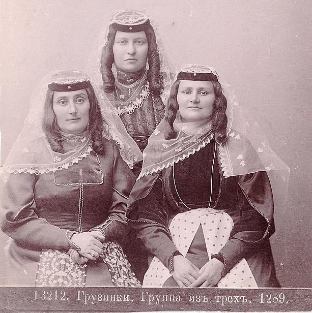 Drei Frauen in traditionellen russischen Hochzeitskleidern mit farbenfroher Stickerei und Perlen stehen vor einer weißen Wand in einem Schwarz-Weiß-Foto.
