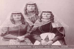 Drei Frauen in traditionellen russischen Hochzeitskleidern mit farbenfroher Stickerei und Perlen stehen vor einer weißen Wand in einem Schwarz-Weiß-Foto.