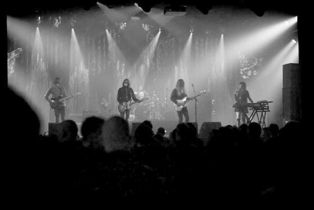 Ein Schwarz-Weiß-Bild eines Musikkonzerts mit vier Künstlern auf der Bühne, darunter drei Gitarristen und ein Sänger, sowie Zuschauern unten und Lichtern oben.