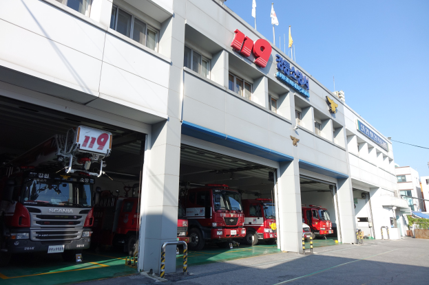 Feuerwache mit mehreren geparkten Feuerwehrautos, einem Gebäude mit Fenstern, Säulen und Namensschildern und Flaggen im Vordergrund vor dem Hintergrund anderer Gebäude und eines klaren blauen Himmels.