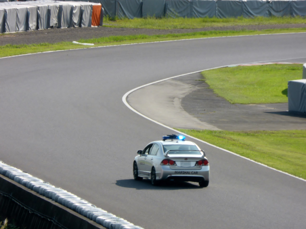 Ein Polizeiauto fährt auf einer Rennstrecke mit Gras auf beiden Seiten, umgeben von Menschen, Zelten und anderen Gegenständen im Hintergrund.